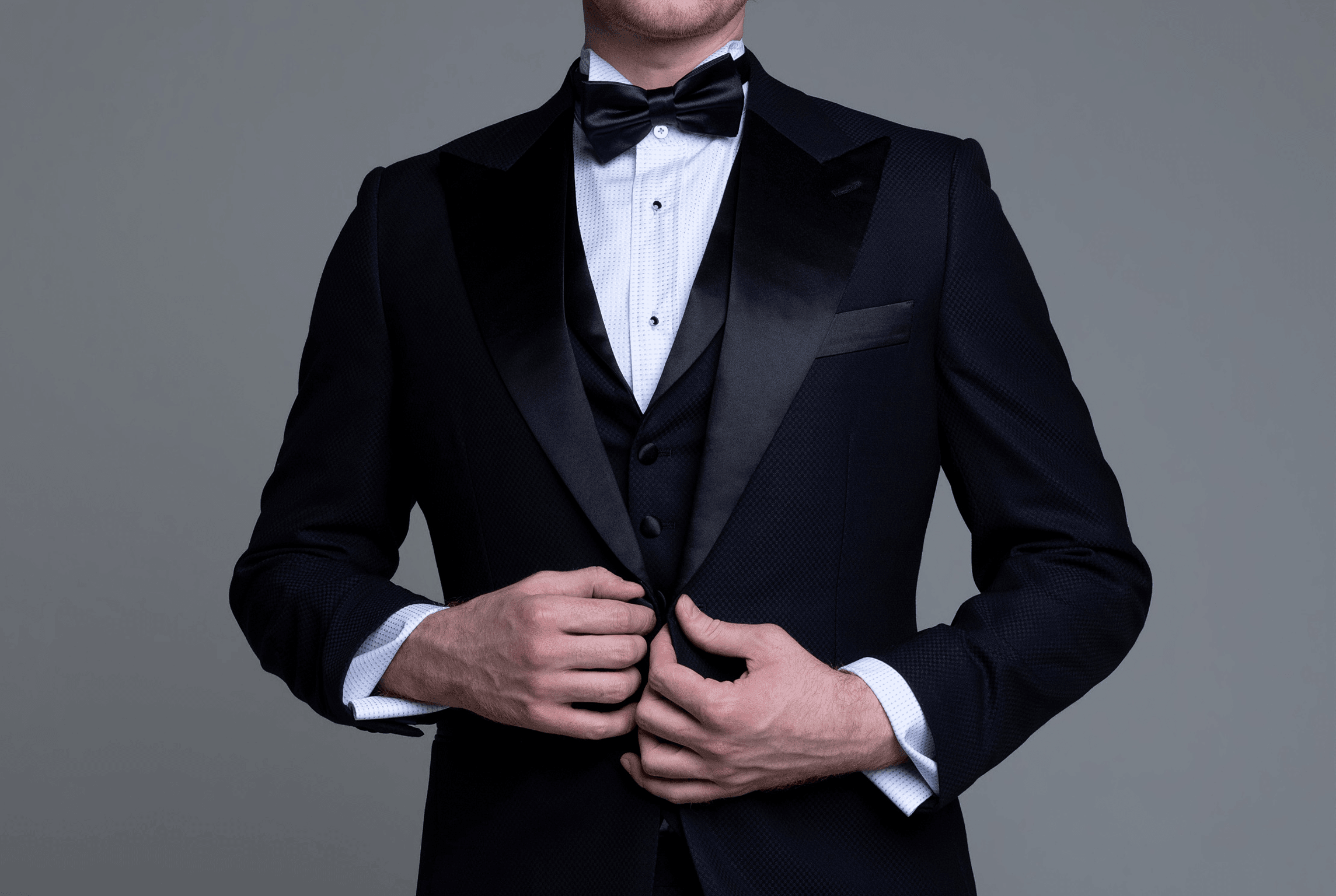 Man in a black tuxedo with satin lapels and bow tie fastening his jacket against a gray studio background.