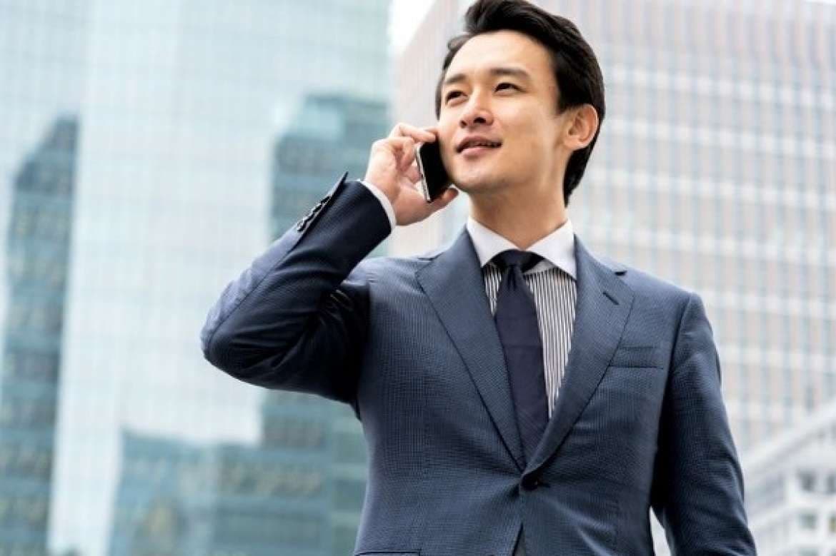 Businessman in a navy suit talking on a phone in front of modern office buildings.