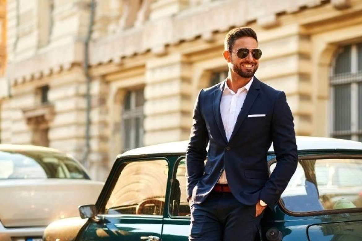 Man in a navy suit and sunglasses standing beside a car.