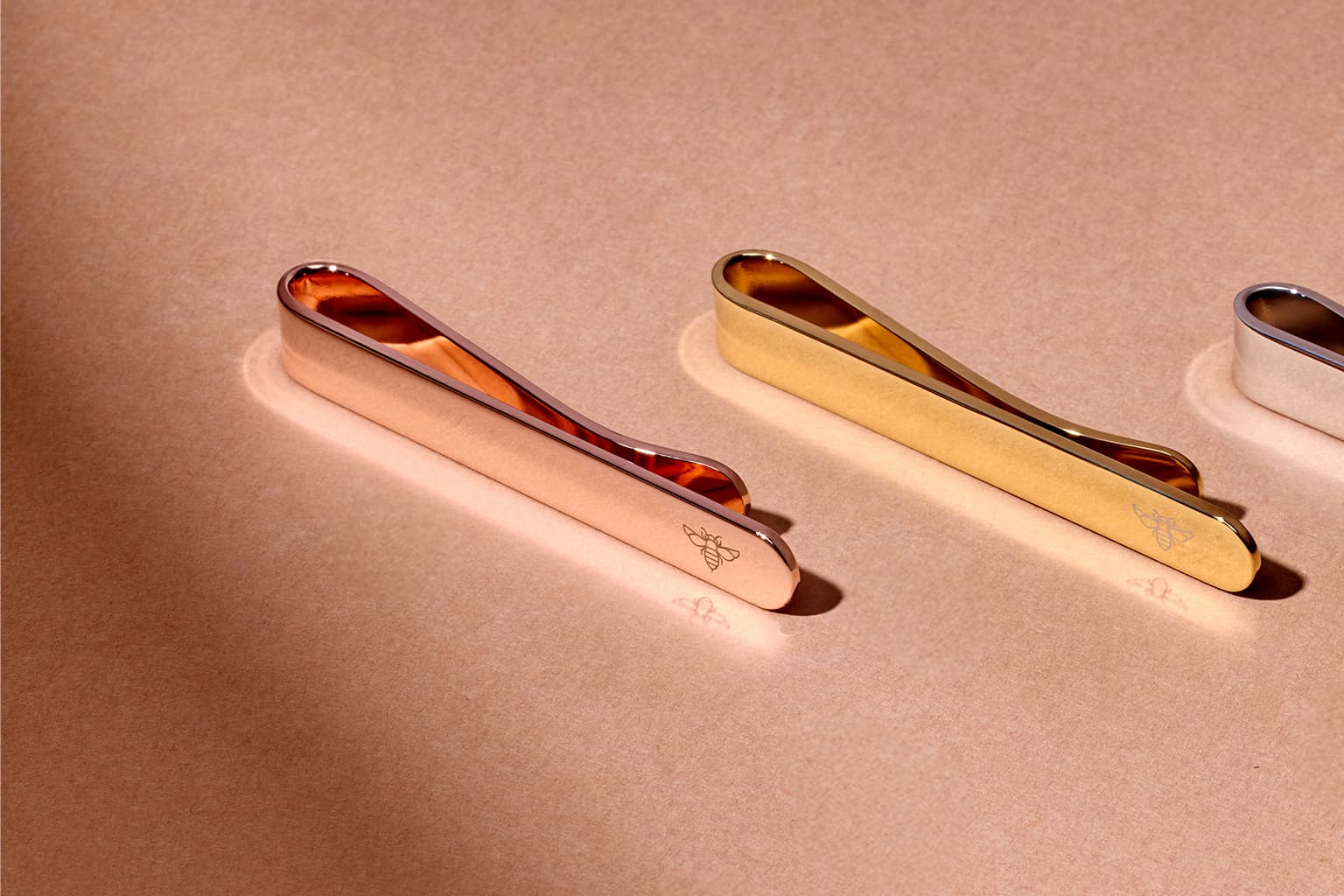 Rose gold, gold, and silver tie bars on a warm surface.