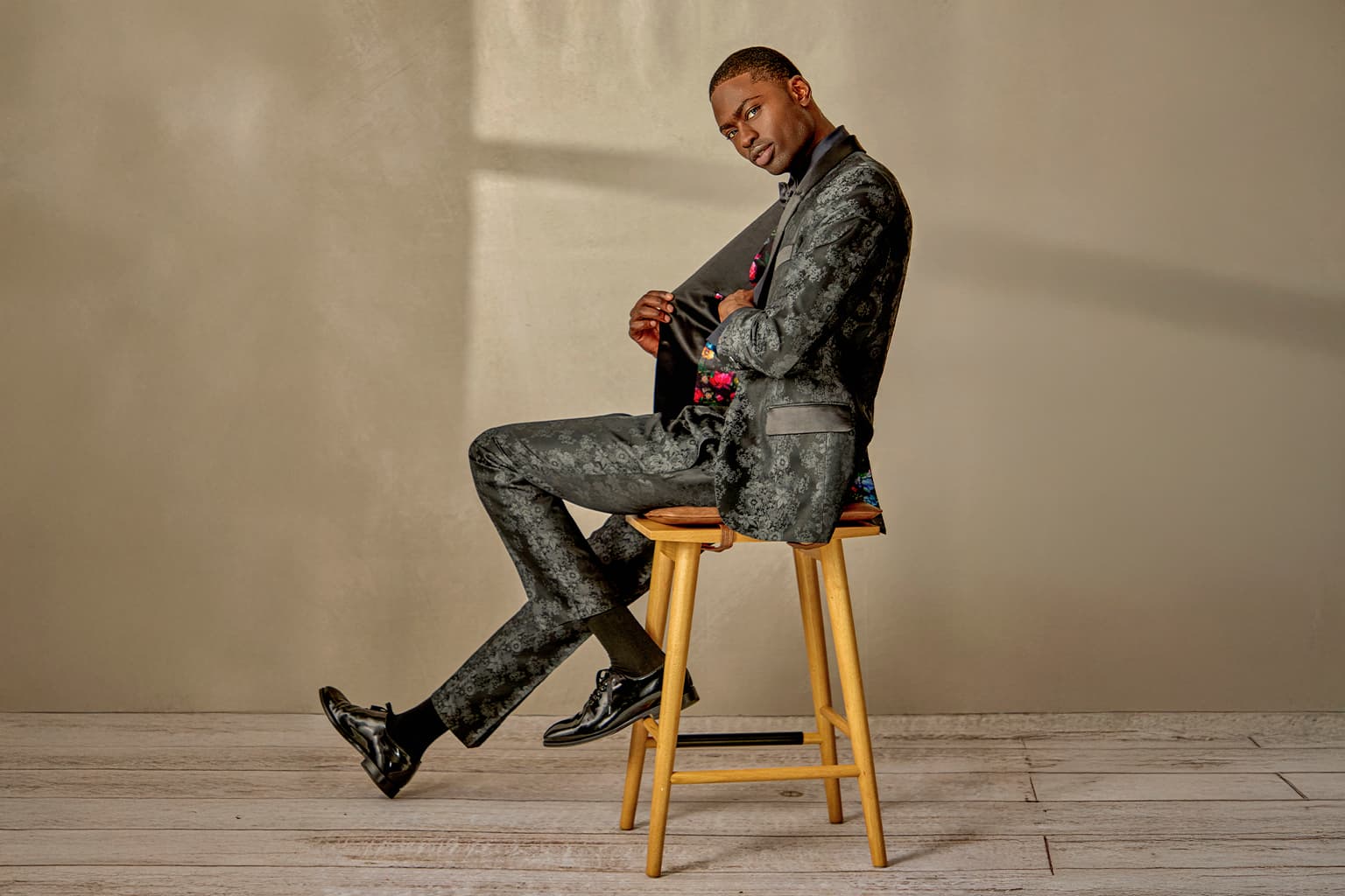 Man seated on a stool in a gray patterned suit.