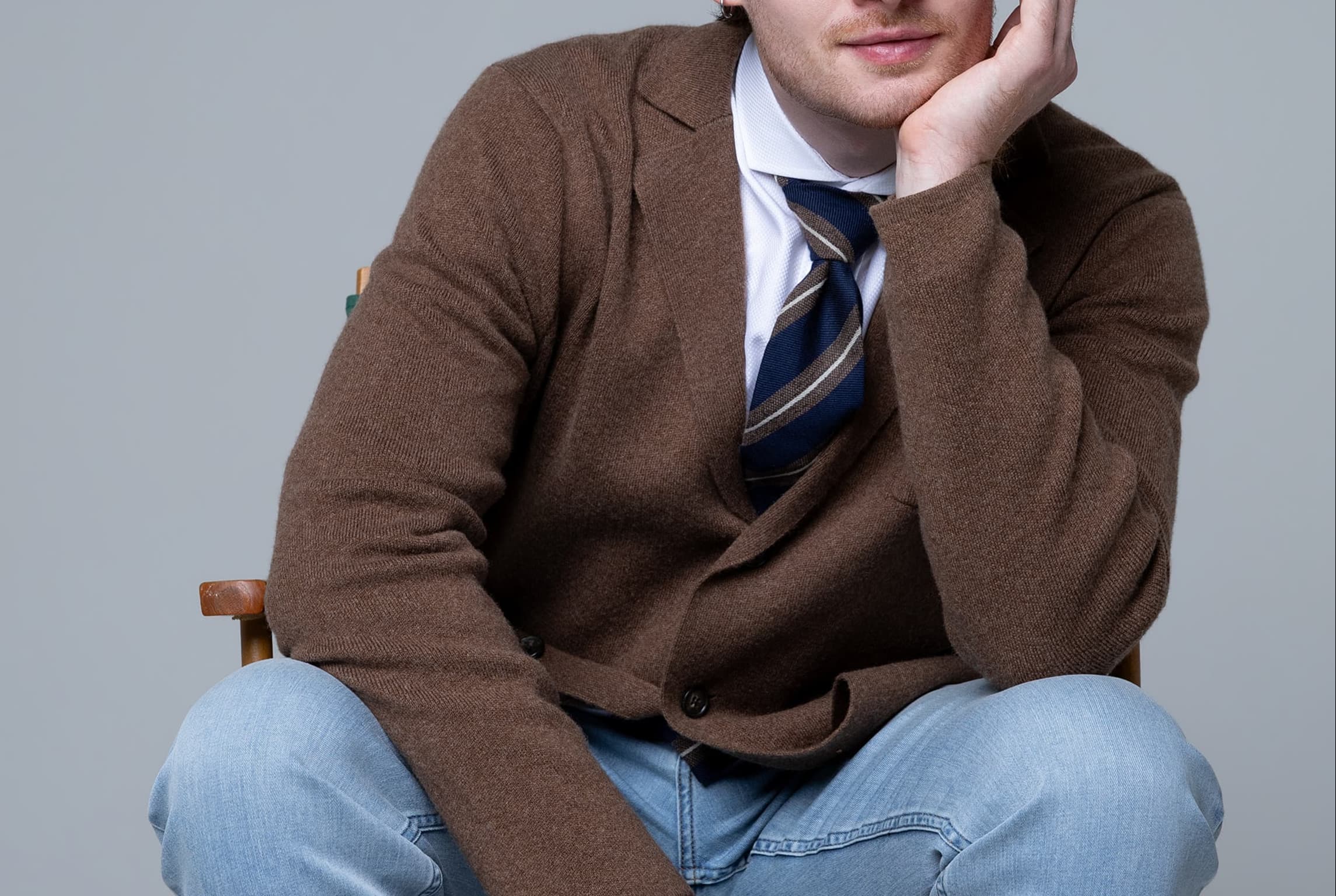 Man wearing a brown cardigan, striped tie, and light-wash jeans while seated against a gray background