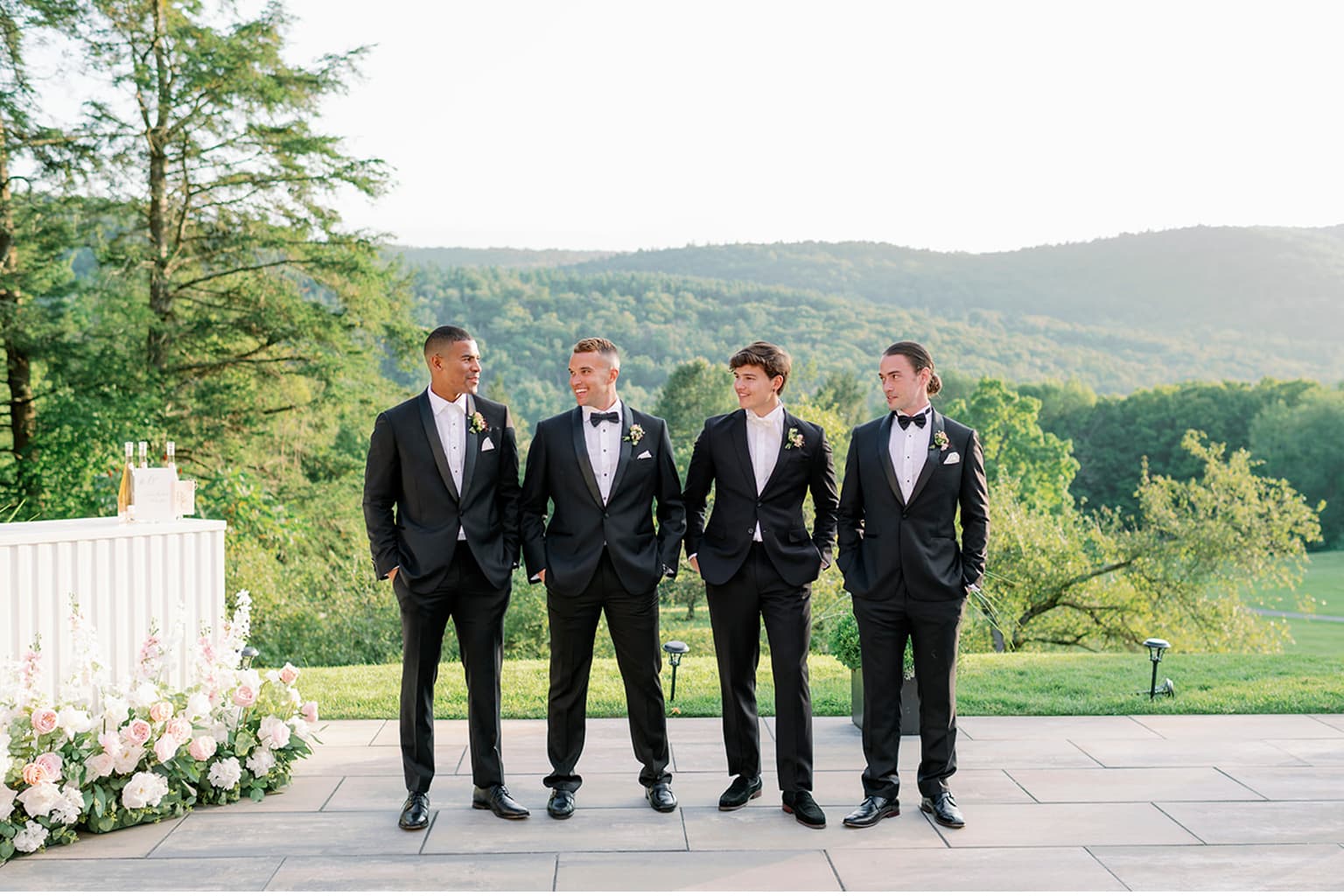 Four groomsmen in black tuxedos standing outdoors at a wedding.