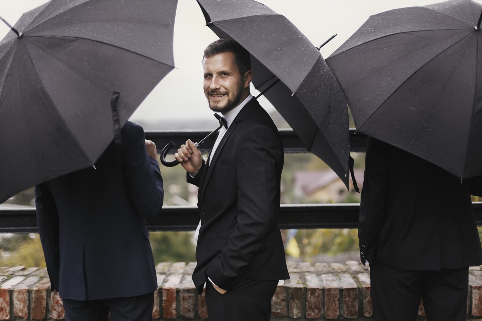 Groom in a black tuxedo standing under umbrellas in the rain.